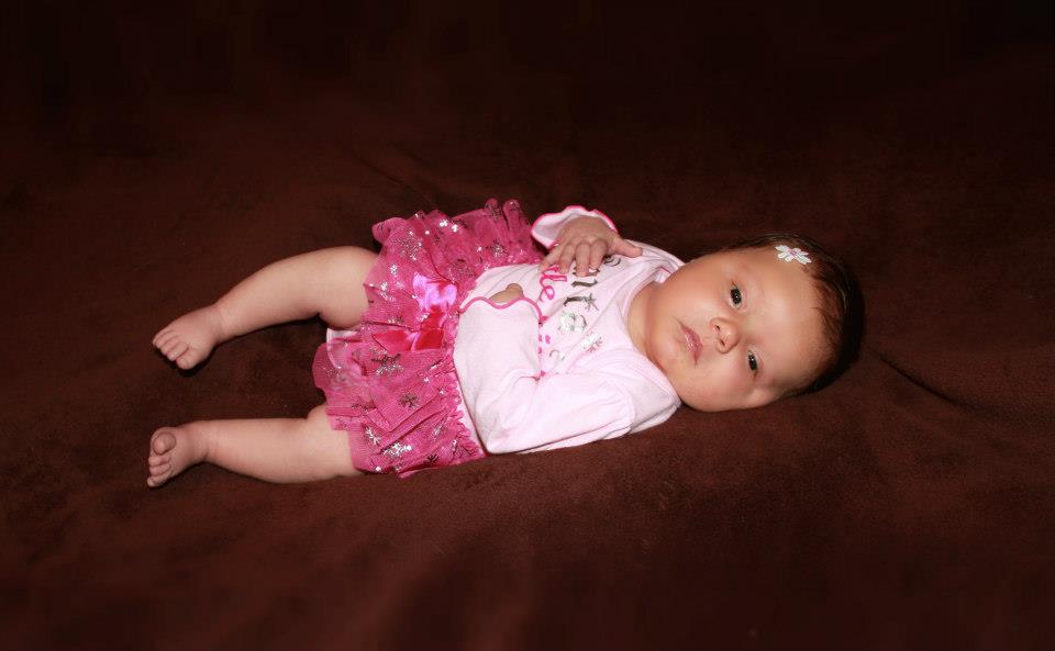 Image description: My one month old daughter in a pink shirt and a tutu against a brown background.